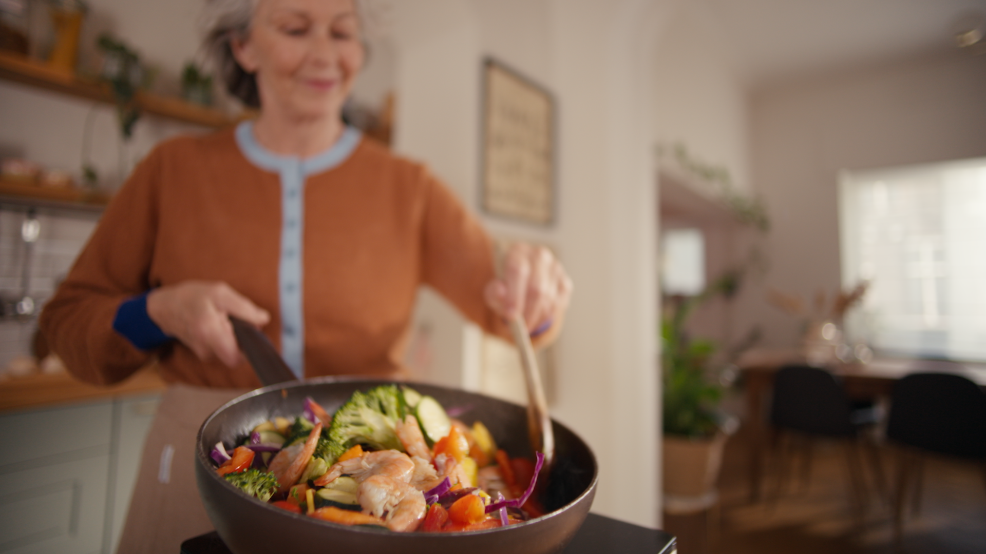 Uśmiechnięta kobieta podrzuca na patelni smażone w ruchu krewetki i wypełnia kuchnię zapachem potraw