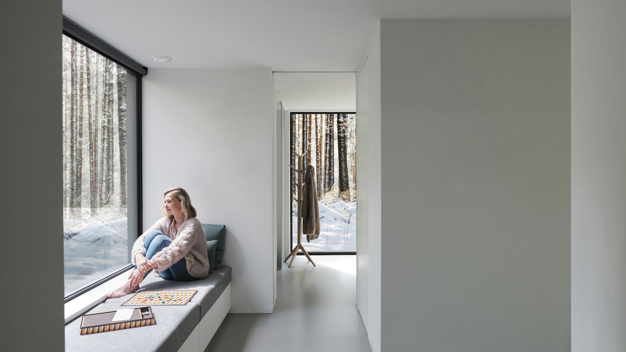 Szerokie ujęcie kobiety siedzącej boso przed oknem i patrzącej na drzewa i śnieg.