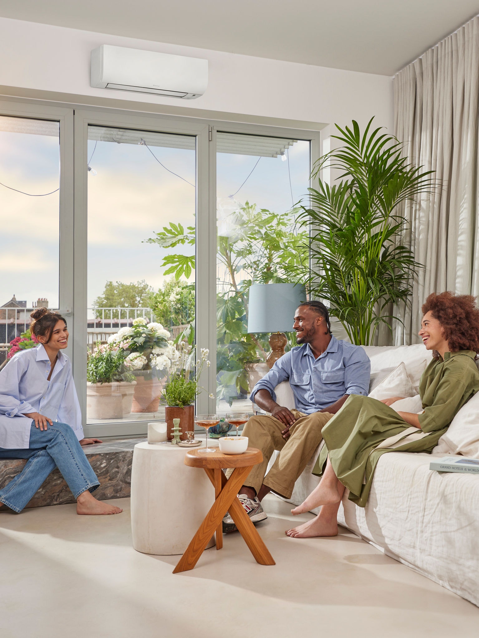 Mężczyzna i dwie kobiety siedzą i rozmawiają w słoneczny dzień w jasnym, przestronnym pokoju z białymi ścianami i dyskretną, białą naścienną pompą ciepła nad dużym oknem