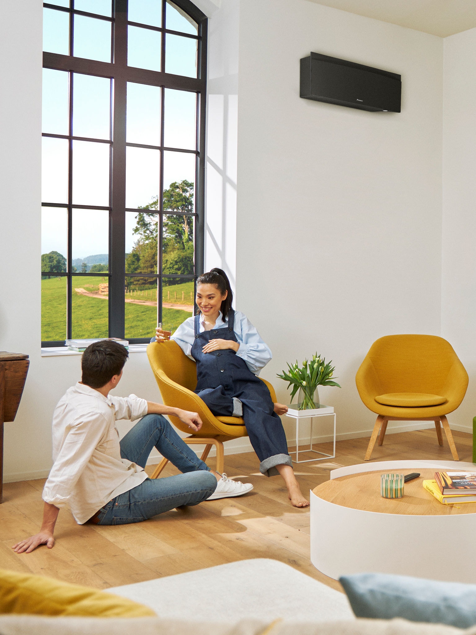 Mężczyzna i kobieta siedzący przed dużym oknem w jasnym pokoju, z czarną pompą ciepła zamontowaną na ścianie za nimi