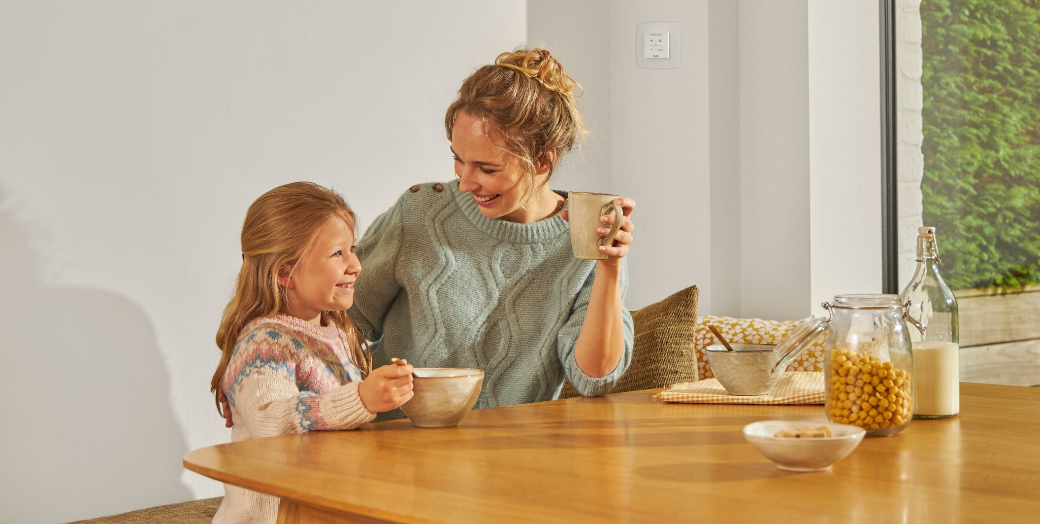 Młoda dziewczyna jedząca z miski siedząca przy stole obok kobiety z kubkiem w ręku, obie uśmiechnięte, szczęśliwe i zrelaksowane, cieszące się zdrowym powietrzem.