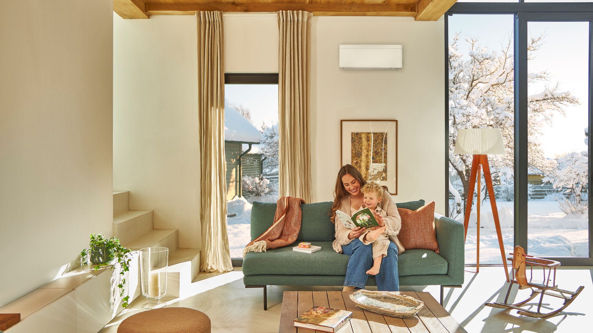 Matka siedząca na kanapie pod pompą ciepła powietrze-powietrze zamontowaną na jednostce naściennej, czytająca książkę dla dzieci z maluchem na kolanach i ciesząca się ciepłem, podczas gdy na zewnątrz pada śnieg.